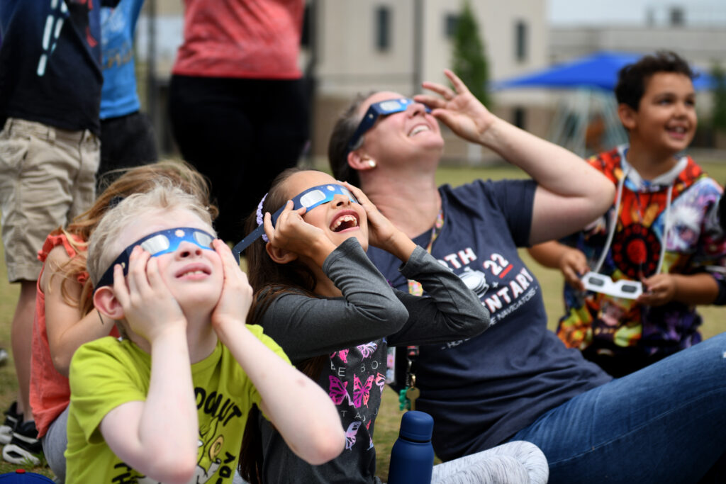 Students and a teacher wearing safety glasses stare up at the eclipse event