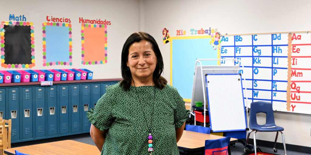 Milagros Noa, a dual-language kindergarten teacher at Whitestone Elementary School, stands in her classroom and smiles for a portrait. In the background, lockers, a whiteboard, and other classroom items are visible.
