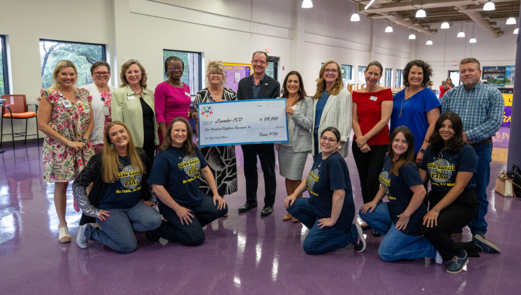 Group of people posing with a large ceremonial check from the Texas PTA to Leander ISD. Some are standing while others are kneeling in the front, all smiling in a brightly lit room.