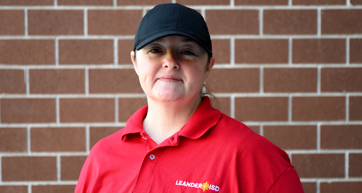 Person in a red Leander ISD polo shirt and black cap, standing in front of a brick wall.